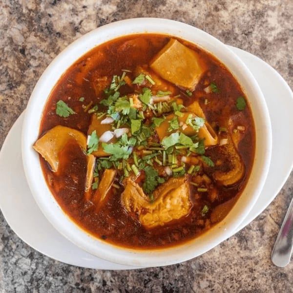 Menudo soup in a white bowl, garnished with cilantro and diced onions, from La Tiendita Mexican Restaurant.
