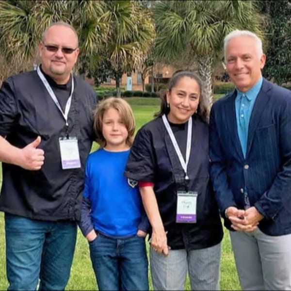 Four people, two men, a woman, and a child, stand smiling outdoors in front of palm trees.