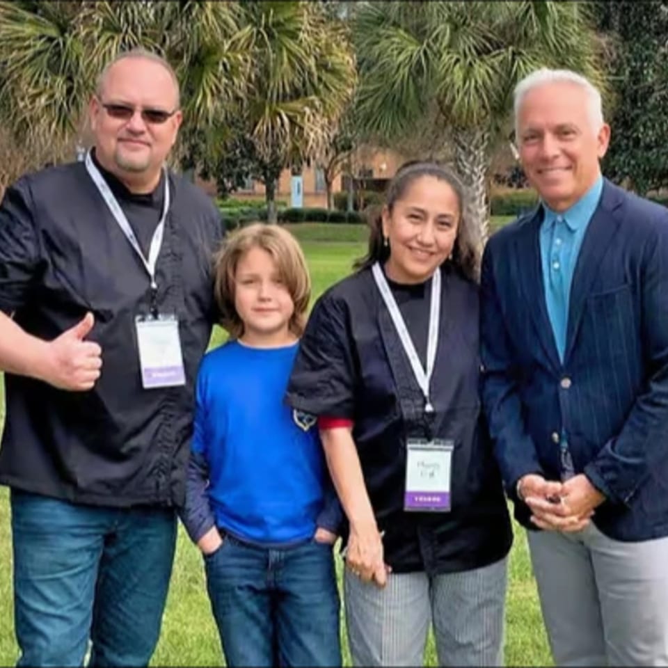 Four people, two men, a woman, and a child, stand smiling outdoors in front of palm trees.