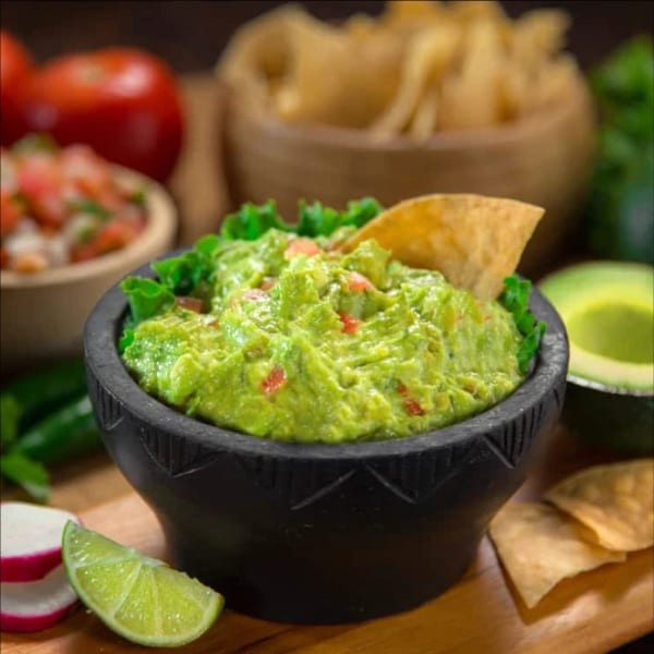 Fresh guacamole in a black bowl with a tortilla chip, lime, radishes, and salsa.