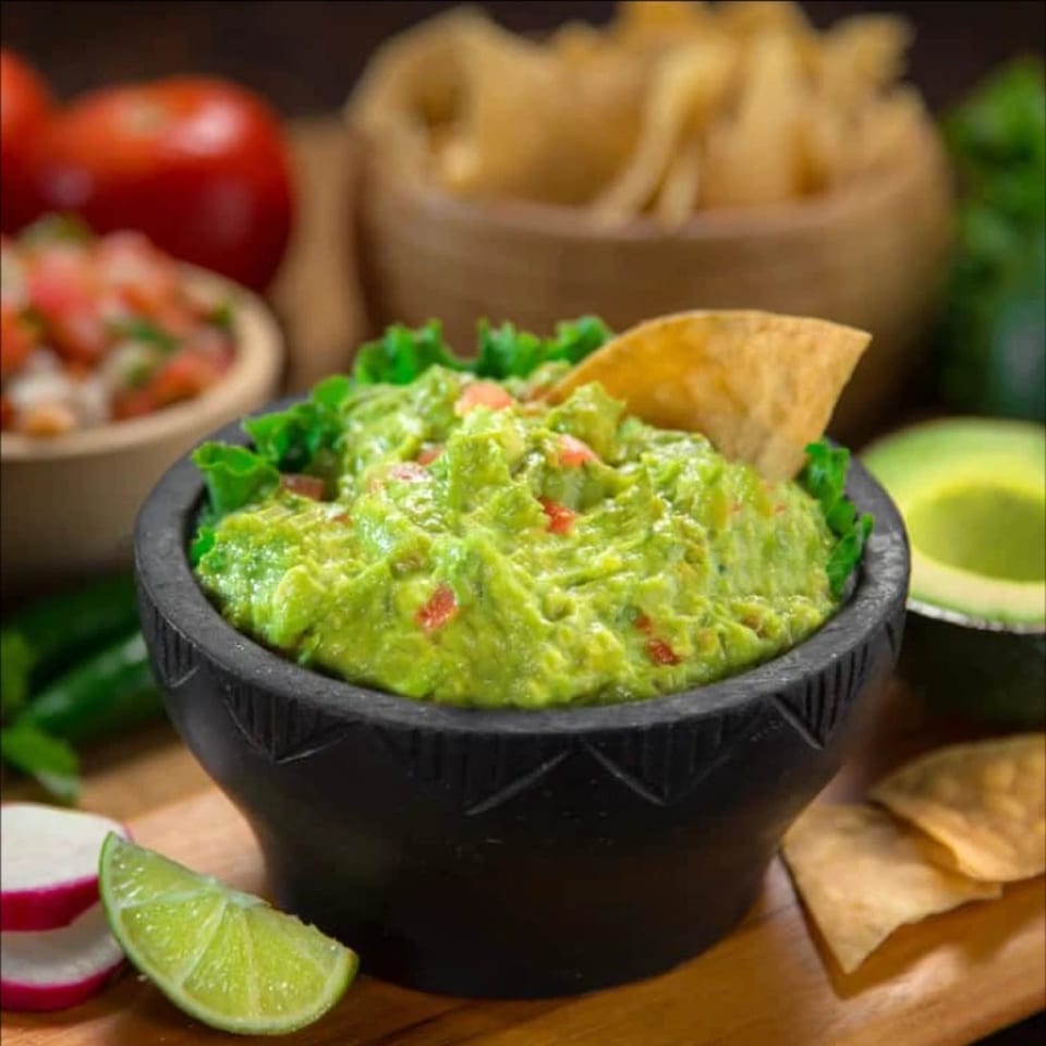 Fresh guacamole in a black bowl with a tortilla chip, lime, radishes, and salsa.